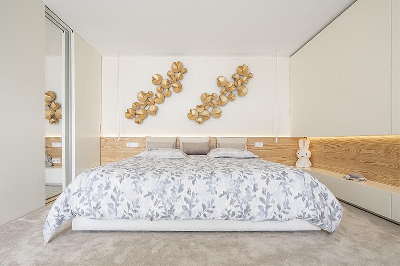 The wood accent adds a natural element to this modern bedroom and creates a headboard for the bed, while between the cabinets to the right of the bed, has hidden lighting, adding a soft glow to the room and highlighting the displayed objects on the shelf. #BedroomDesign #ModernBedroom #WoodAccent, #Lighting