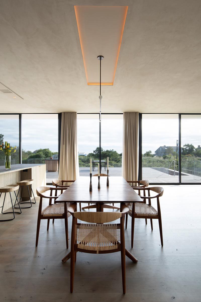 In this modern dining area, there's a recess in the ceiling with hidden lighting, while a minimalist light hangs above a wood dining table. Sliding glass doors open to the deck outside that has ocean views. #DiningRoom #Lighting #ModernInterior