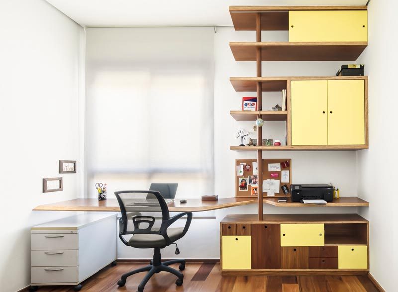 In this modern home office, there's a custom-designed desk that fills the width of the wall, and a wood shelving unit that adds a soft pop of color with yellow doors. #HomeOffice #ModernDesk #Shelving