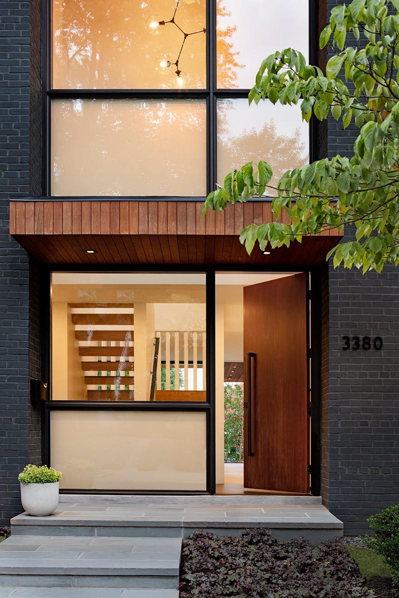 A wood front door welcomes visitors to this modern house and opens to a small light-filled foyer. #ModernFrontDoor #Windows