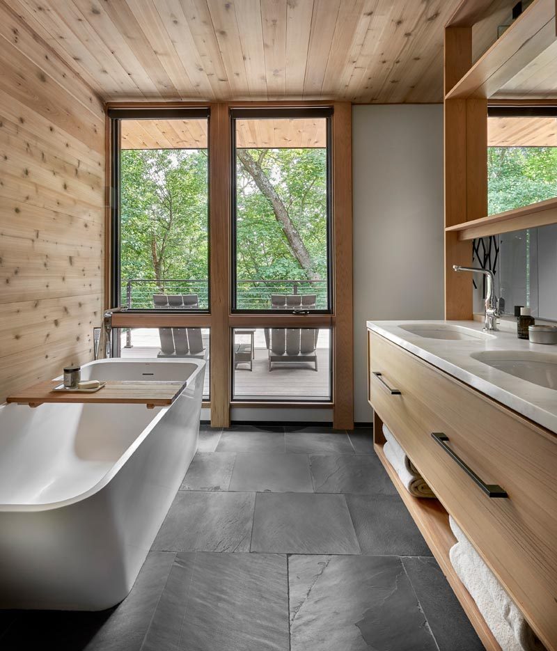 In this modern master bathroom, the freestanding bathtub takes advantage of the windows that overlook a small deck. Opposite the bathtub is a wood vanity with dual sinks, storage drawers, and open shelving. #MasterBathroom #BathroomDesign