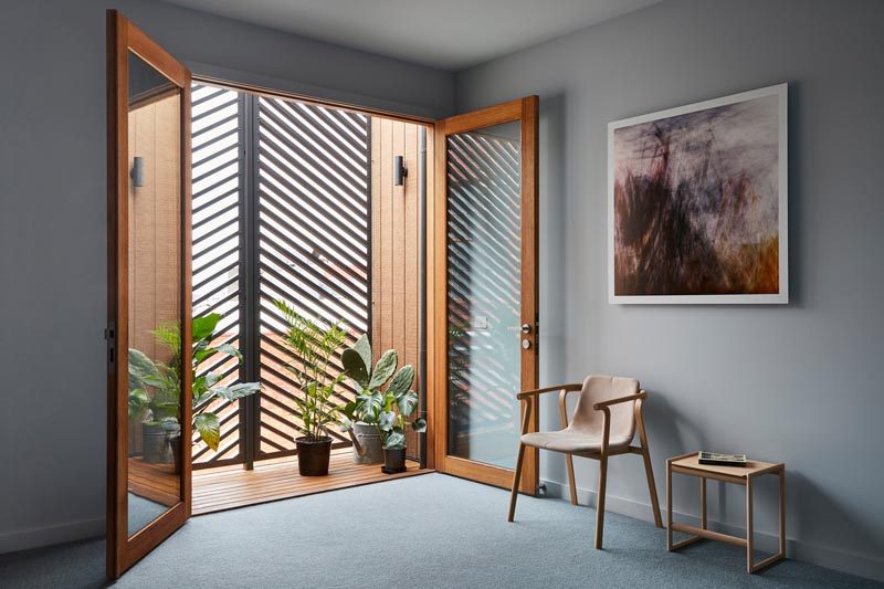 In this modern bedroom with grey walls, wood-framed glass doors open up to a small balcony that hides behind the horizontal slat panels on the facade. #ModernBedroom #GlassDoors #EnclosedBalcony