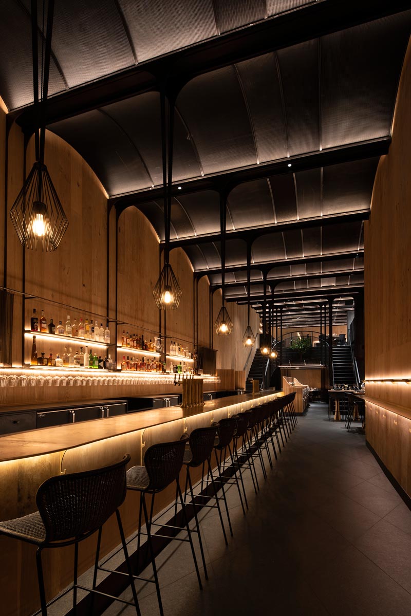 This modern restaurant has a high vaulted ceiling, wood walls, hidden lighting, an expansive bar, and various dining areas, including booth and banquette seating. #RestaurantInterior #ModernRestaurant #BarInterior #BarDesign