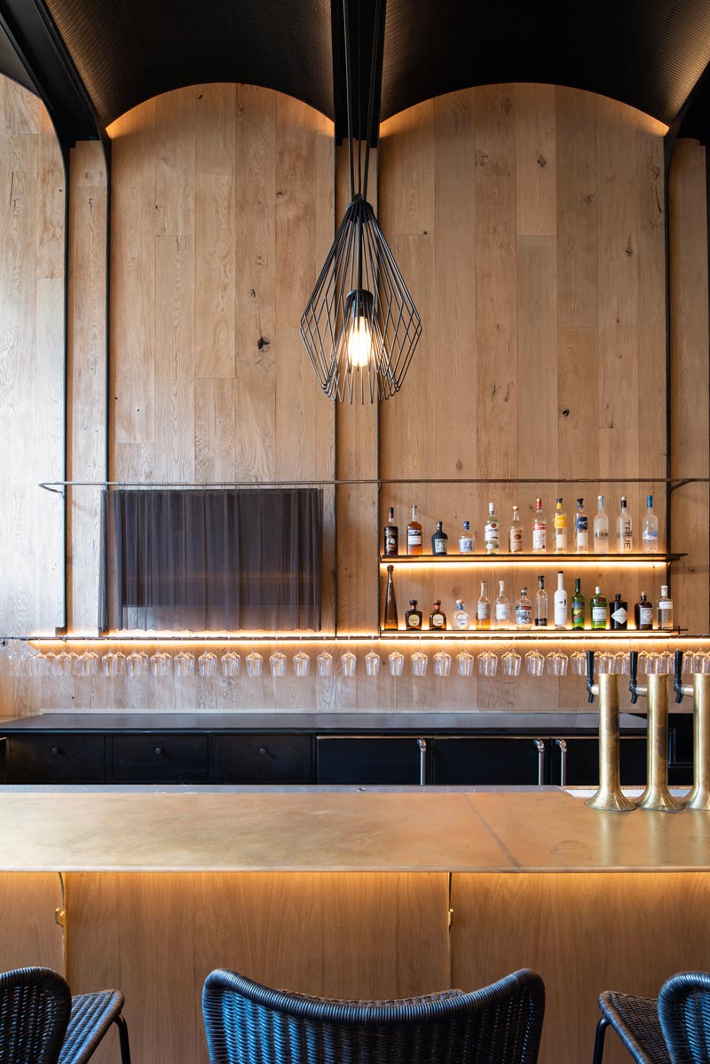This modern restaurant has a high vaulted ceiling, wood walls, hidden lighting, an expansive bar, and various dining areas, including booth and banquette seating. #RestaurantInterior #ModernRestaurant #BarInterior #BarDesign