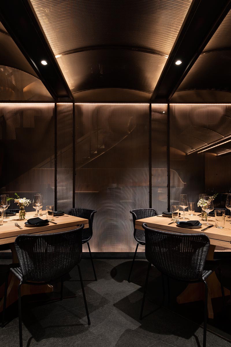 This modern restaurant has a high vaulted ceiling, wood walls, hidden lighting, an expansive bar, eye-catching metal stairs, and various dining areas, including booth and banquette seating. #RestaurantInterior #ModernRestaurant #BarInterior #BarDesign