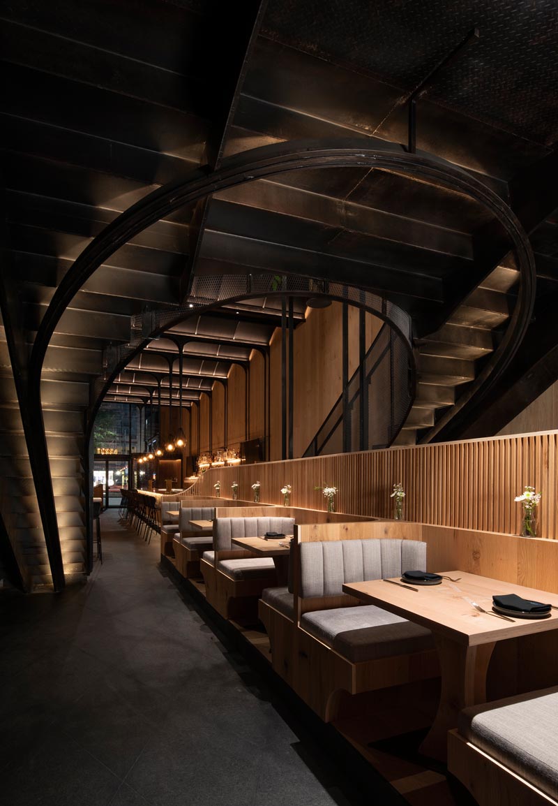 This modern restaurant has a high vaulted ceiling, wood walls, hidden lighting, an expansive bar, and various dining areas, including booth and banquette seating. #RestaurantInterior #ModernRestaurant #BarInterior #BarDesign