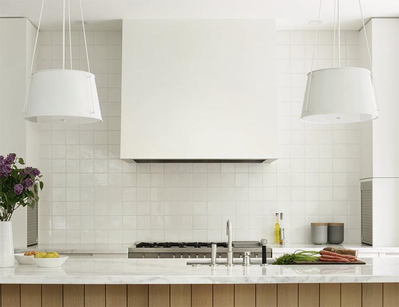 This modern kitchen features two hanging pendant lights, an undermount sink, a white tile backsplash, stainless steel appliances, and a range hood that matches the cabinets. #ModernKitchen #KitchenDesign #WhiteKitchen