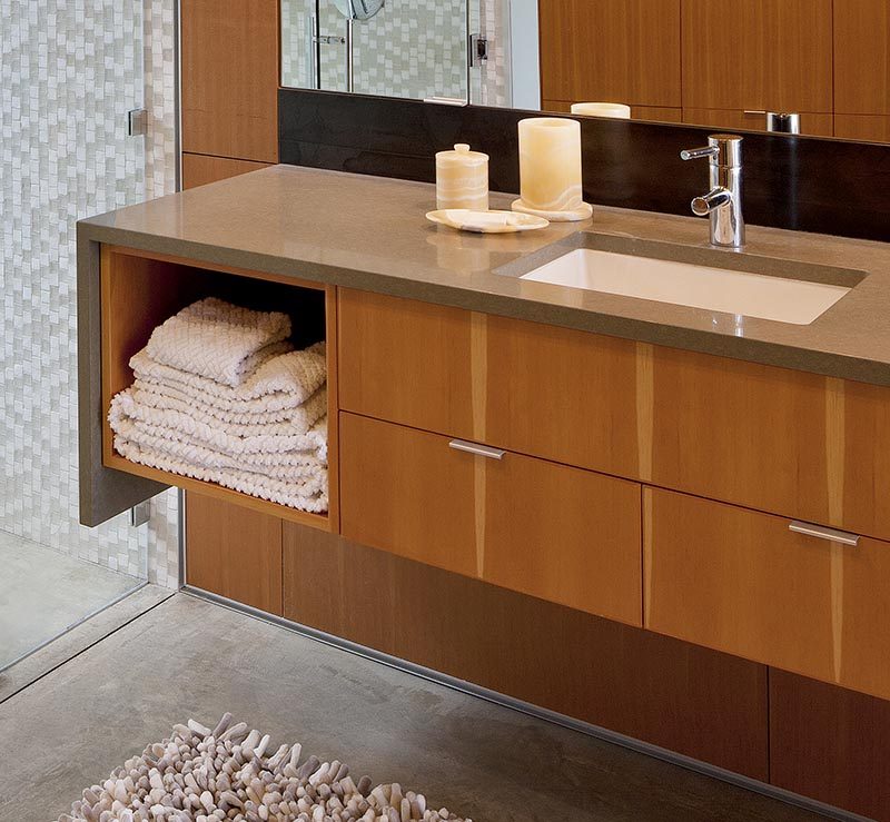 This modern bathroom has a floating wood vanity that features an open shelf and drawers, a waterfall edge counter made from Ceasarstone in mocha with a small fleck pattern, an undermount sink, and a Hansgrohe faucet. #ModernBathtub #FloatingVanity #WoodVanity #Ceasarstone