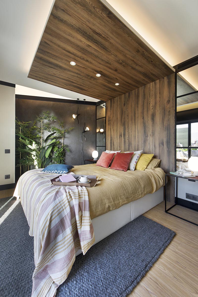 This modern bedroom has a wood headboard that wraps from the wall up onto the ceiling, and acts as an open wood canopy adding a natural touch to the room and defining an area for the bed. It also has hidden lighting that creates a soft glow and highlights the design element. #WoodHeadboard #WrapAroundHeadboard #ModernBedroom #BedroomDesign