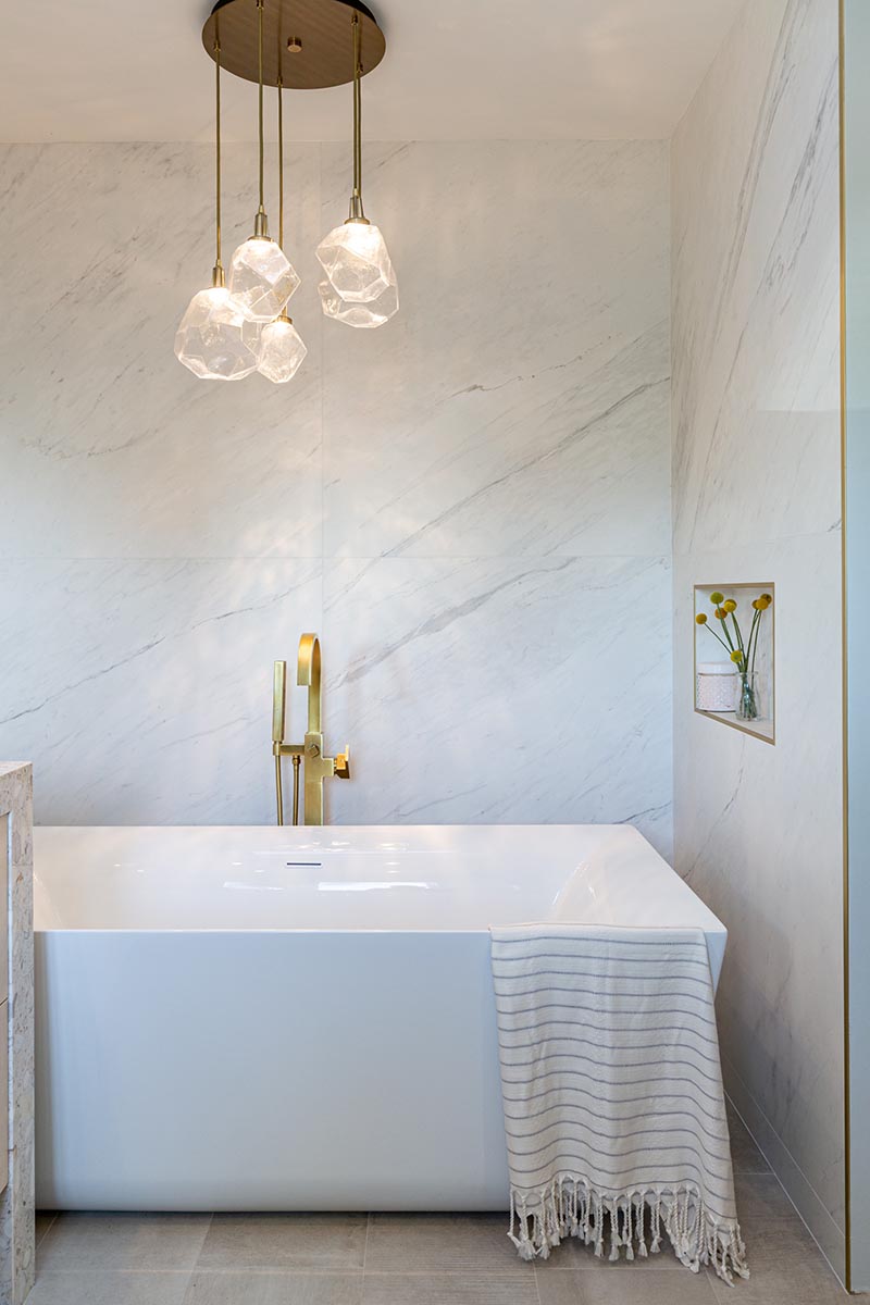 This modern bathroom has a freestanding white bathtub that's recessed into its own alcove. A sculptural multi-light pendant lamps with glass geometric shades, hangs above the bath, highlighting the design. #Bathtub #ModernBathroom