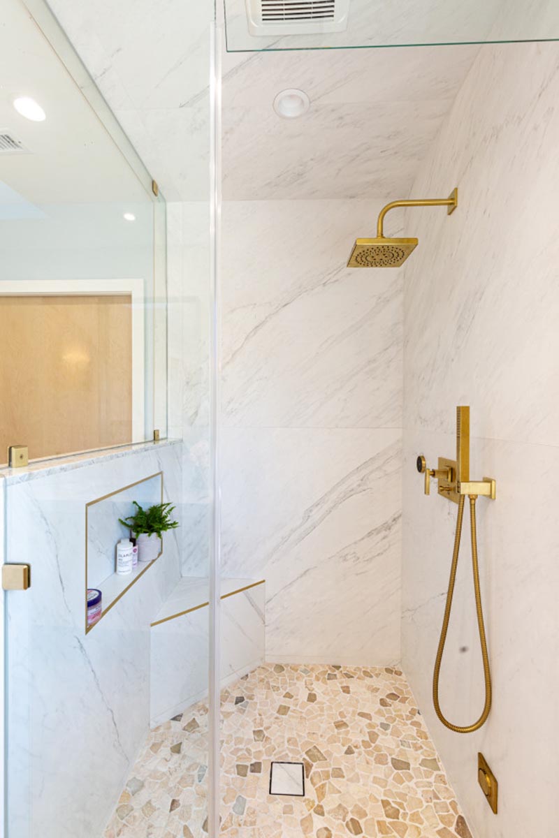 Inside this modern shower, the brass shower head and shower wand contrast the neutral stone, while a shower niche with a brass border provides a place for storage. A small corner bench has also been included in the design of the shower. #ModernShower #GlassShower #EnclosedShower #BrassAccents