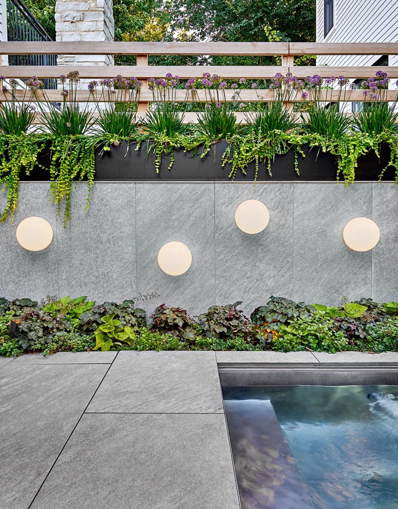 This modern outdoor space features a stone-look porcelain tile, while native, drought-resistant plantings add a natural touch, and globe light fixtures add an artistic flair to the wall by the custom-designed spa. #Landscaping #BlackSteelPlanters #OutdoorLighting #OutdoorEntertaining