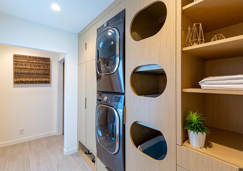 Laundry Ideas - This modern laundry room is designed with separate cubbies for sorting clothes, built-in washer and dryer, the pull-out step located below them, plenty of storage space, open shelving, and a pet feeding area. #LaundrySorter #LaundryDesign #ClothesSorter #BuiltInWasherDryer #ModernLaundry