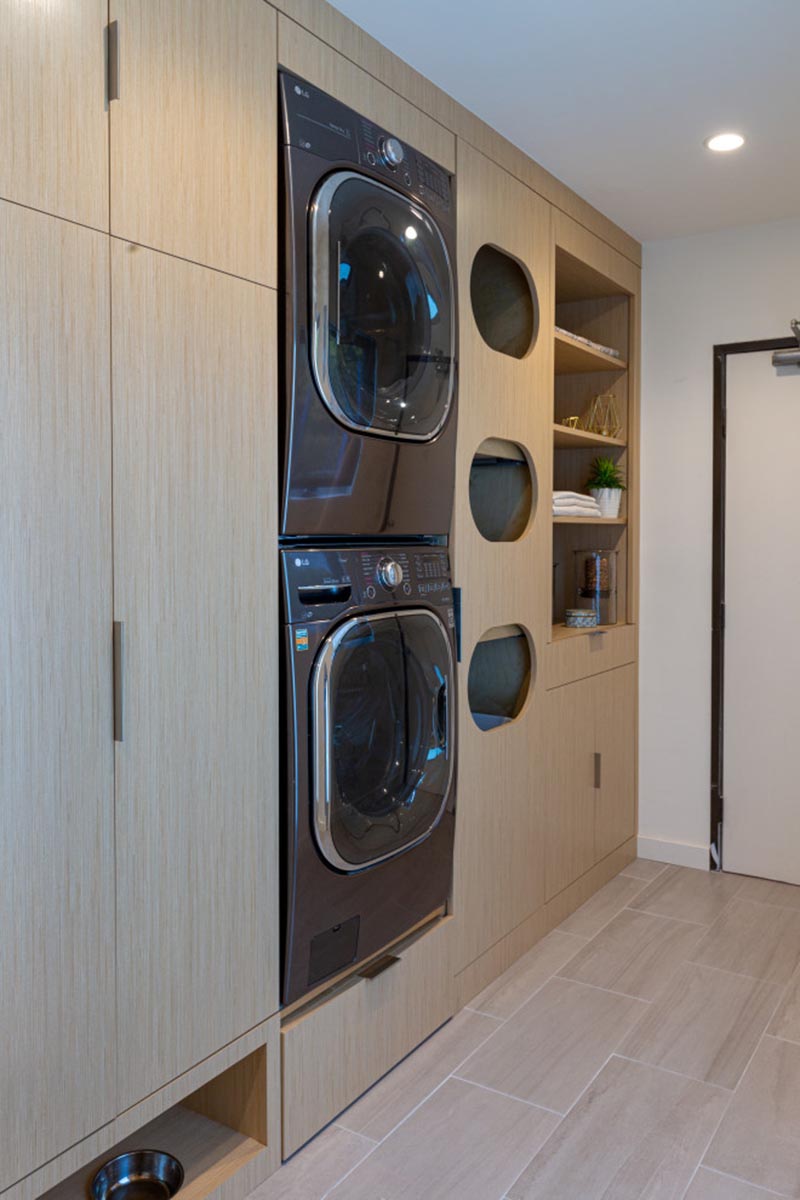 Laundry Ideas - This modern laundry room is designed with separate cubbies for sorting clothes, built-in washer and dryer, the pull-out step located below them, plenty of storage space, open shelving, and a pet feeding area. #LaundrySorter #LaundryDesign #ClothesSorter #BuiltInWasherDryer #ModernLaundry