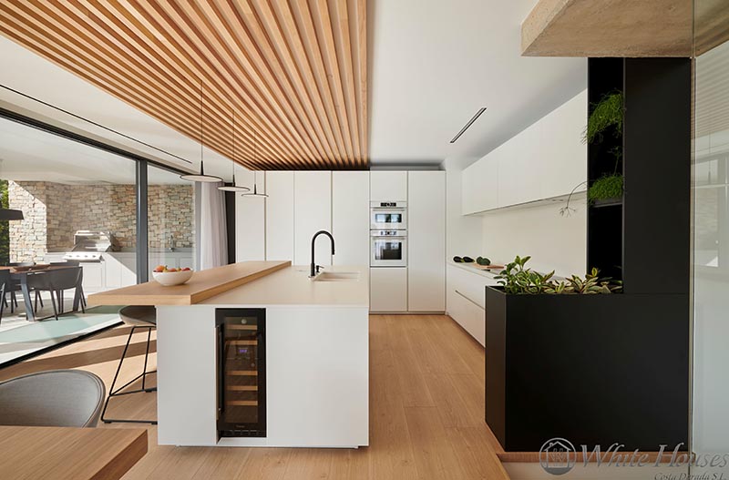 In this modern kitchen, the island has a small built-in wine fridge, while black planter at the end of the cabinets, travels up to the ceiling with shelves for even more plants, adding a contrasting element and a natural touch. #Kitchen #ModernKitchen #WineFrige #Planter