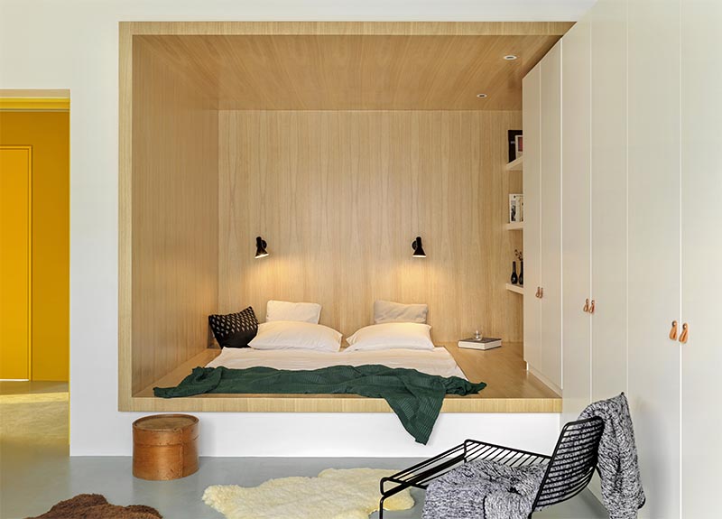 Instead of having a freestanding bed, the designers made use of an odd  shaped corner small positioned the bed within the base of its own wood-lined niche, allowing for more floor space by the large windows. #BedroomIdea #BedNook #BedNiche #BuiltInBed #InteriorDesign #ModernBedroom