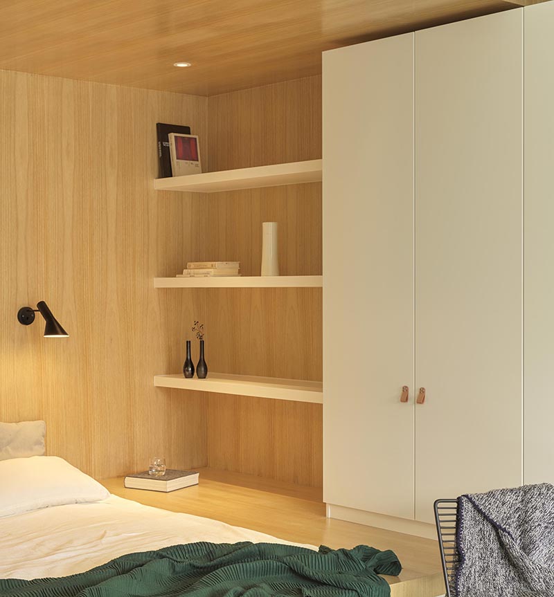 Instead of having a freestanding bed, the designers made use of an odd  shaped corner small positioned the bed within the base of its own wood-lined niche, allowing for more floor space by the large windows. #BedroomIdea #BedNook #BedNiche #BuiltInBed #InteriorDesign #ModernBedroom #Shelving