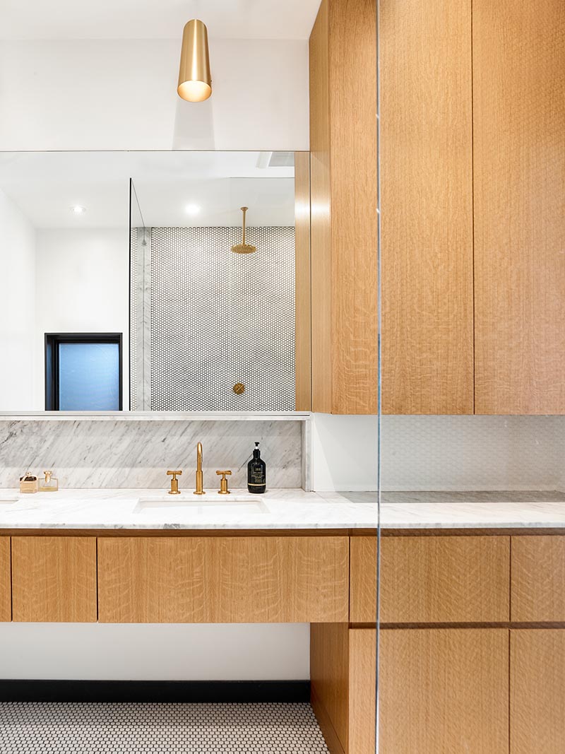 A modern master bathroom with hardware free wood cabinets, a marble vanity with dual sinks, brass fixtures, penny tiles, and custom-build niches. #ModernBathroom #MarbleVanity #BrassFixtures #PennyTiles #ShowerNiche