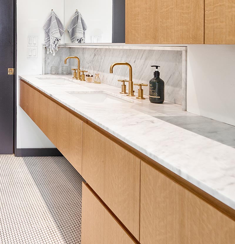 For the vanity in this modern bathroom, hardware free wood cabinets have been paired with light grey marble countertops, white under-mount dual sinks, and brass fixtures. #ModernBathroomVanity #MarbleVanity #BrassFixtures #WoodCabinets