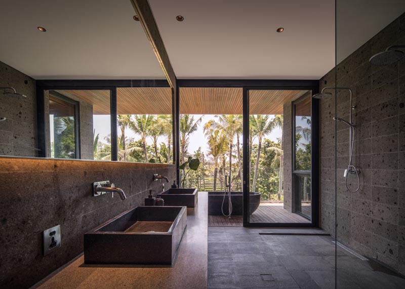 In this modern bathroom, the freestanding bathtub is located on the covered balcony, while inside, hidden lighting behind the mirror highlights the stone wall and vanity. #ModernBathroom #BathroomDesign #OutdoorBathtub