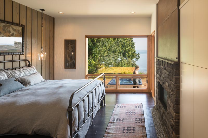 This master bedroom has a fireplace and a large wood-framed window that looks out to the water. #BedroomDesign