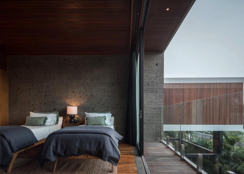 In this modern bedroom, the stone walls provide a earthy grey backdrop for the beds, while a sliding glass door opens to a small balcony with a glass railing, that provides uninterrupted views of the treetops and the outdoor space below. #ModernBedroom #Sandstone #BedroomDesign #Balcony