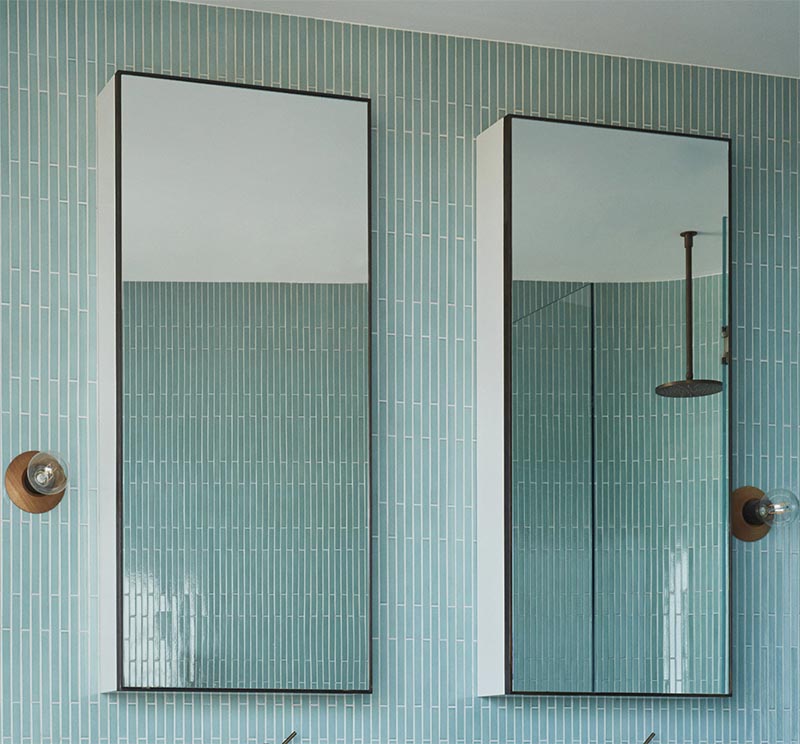 Above the vanity in this modern bathroom is a duo of vertical black framed mirrors. Adjacent to each mirror is a simple sconce with a wood base and an exposed bulb. #BathroomMirrors #ModernBathroom #BlueTIles #BathroomDesign