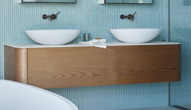 This modern bathroom has a floating wood vanity with curved corners, adding to the softness of the space, while a thin white countertop is home to a pair of minimalist white vessel sinks. #FloatingVanity #WoodVanity #ModernBathroom