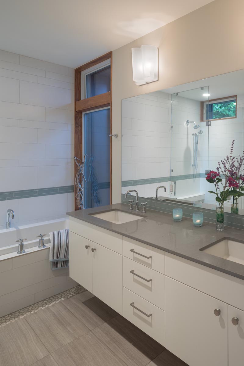 In this modern bathroom, a white vanity with double sinks is topped with a grey countertop, while white tiles adjacent to the bathtub have a strip of color. #BathroomDesign #WhiteBathroom