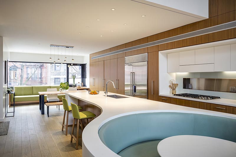 The custom-designed white Corian island countertop in this modern kitchen, has a rectangular shape at one end, before transitioning into a curved split design that wraps around the soft blue banquette seating that makes up the integrated breakfast nook. #KitchenDesign #ModernKitchen #BreakfastNook #BanquetteSeating #KitchenIsland