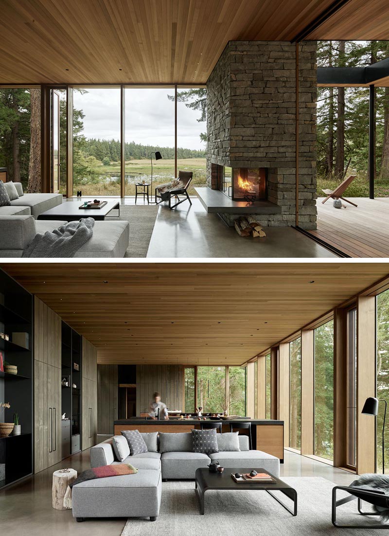 In this modern open plan living room, a stone fireplace with a concrete hearth, and the views of the land are the main focal points. #LivingRoom #Fireplace #Windows #WoodCeiling #ModernHouse