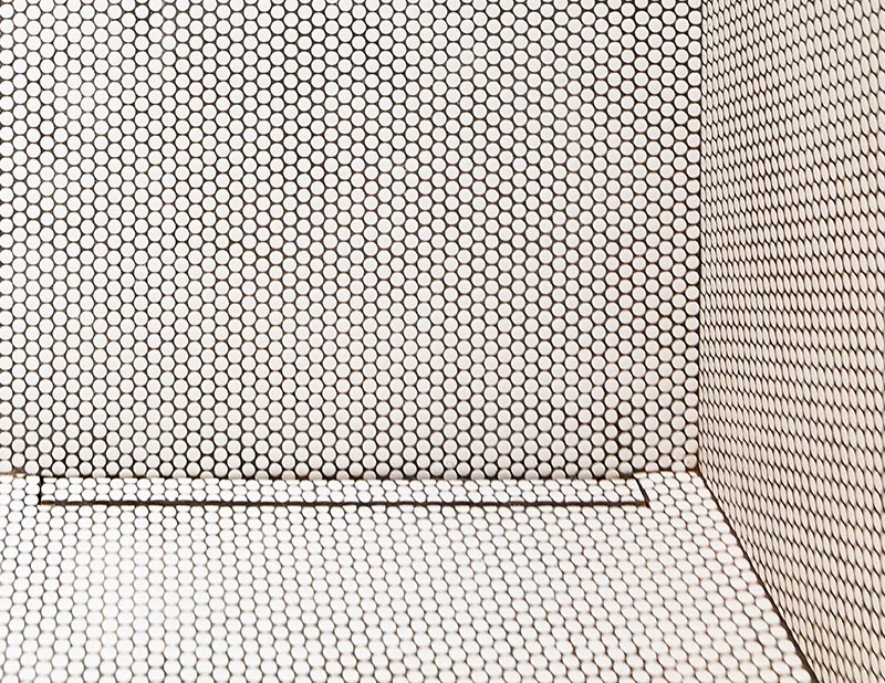 White penny tiles with black grout have been used for both the flooring throughout this modern bathroom and to line the shower stall. In the floor of the shower, there's a linear drain that's also been tiled to match its surroundings, allowing it to almost blend in. #PennyTile #BlackGrout #LinearDrain #ShowerDrain #InvisibleShowerDrain #ModernBathroom #BathroomDesign