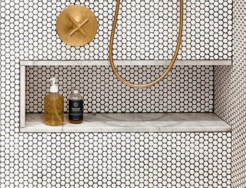 A shower niche lined with the same marble as the vanity has been added in the shower to provide a storage place for the shampoo bottles and soap. This allows the design of the shower to shine through, as there's no additional shelf attached to the wall or hanging from the shower fixtures. #ShowerNiche #PennyTiles #BrassFixtures #MarbleAccent #ModernBathroom