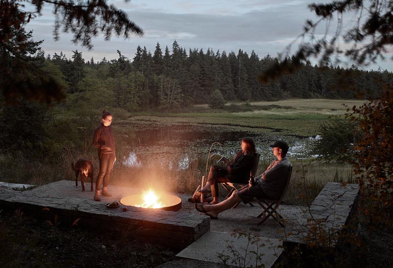 An outdoor entertaining area with a fire pit and low stone walls looks out to a meadow and a pond. #FirePit #OutdoorSpace