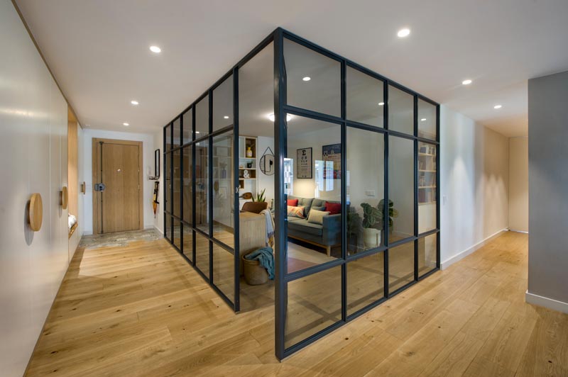 Inside this modern glass-enclosed home office is a long wood desk that meets a custom-designed bookshelf, with some of the shelves lined with wood. Opposite the desk is a comfortable grey couch, a few plants, and colorful pillows, while a rug adds a soft touch underfoot. #HomeOffice #GlassWalls #Shelving