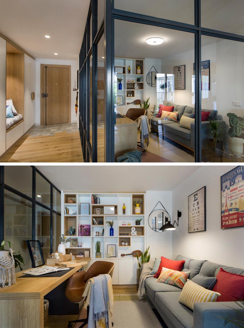 Inside this modern glass-enclosed home office is a long wood desk that meets a custom-designed bookshelf, with some of the shelves lined with wood. Opposite the desk is a comfortable grey couch, a few plants, and colorful pillows, while a rug adds a soft touch underfoot. #HomeOffice #GlassWalls #Shelving