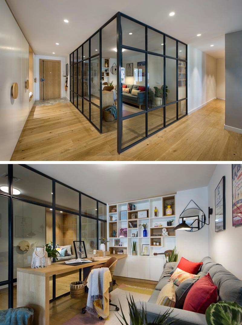 Inside this modern glass-enclosed home office is a long wood desk that meets a custom-designed bookshelf, with some of the shelves lined with wood. Opposite the desk is a comfortable grey couch, a few plants, and colorful pillows, while a rug adds a soft touch underfoot. #HomeOffice #GlassWalls #Shelving