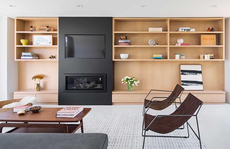 The linear fireplace with a matte black frame is located below a TV that's been recessed into the black accent wall, creating a unified accent in living room that contrasts the light wood. #BuiltInTV #BuiltInTelevision #RecessedTelevision #LinearFireplace #LivingRoomIdeas #Shelving #ModernLivingRoom