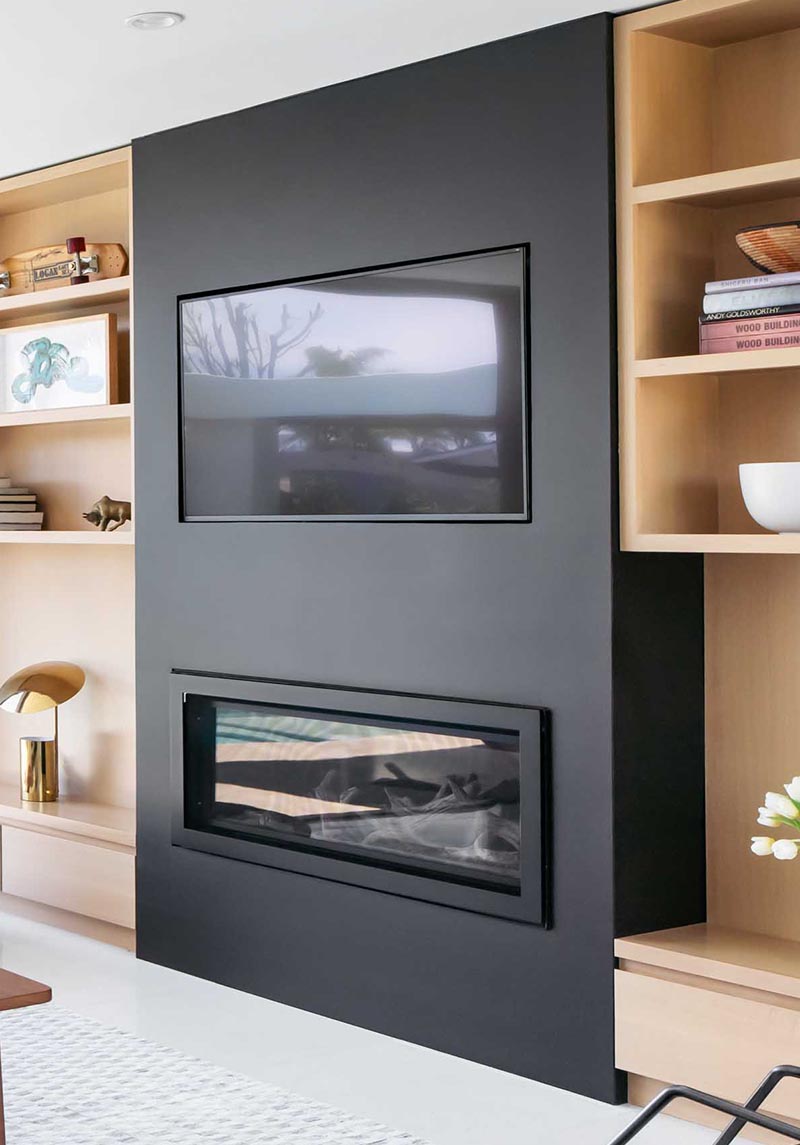The linear fireplace with a matte black frame is located below a TV that's been recessed into the black accent wall, creating a unified accent in living room that contrasts the light wood. #BuiltInTV #BuiltInTelevision #RecessedTelevision #LinearFireplace #LivingRoomIdeas #Shelving #ModernLivingRoom