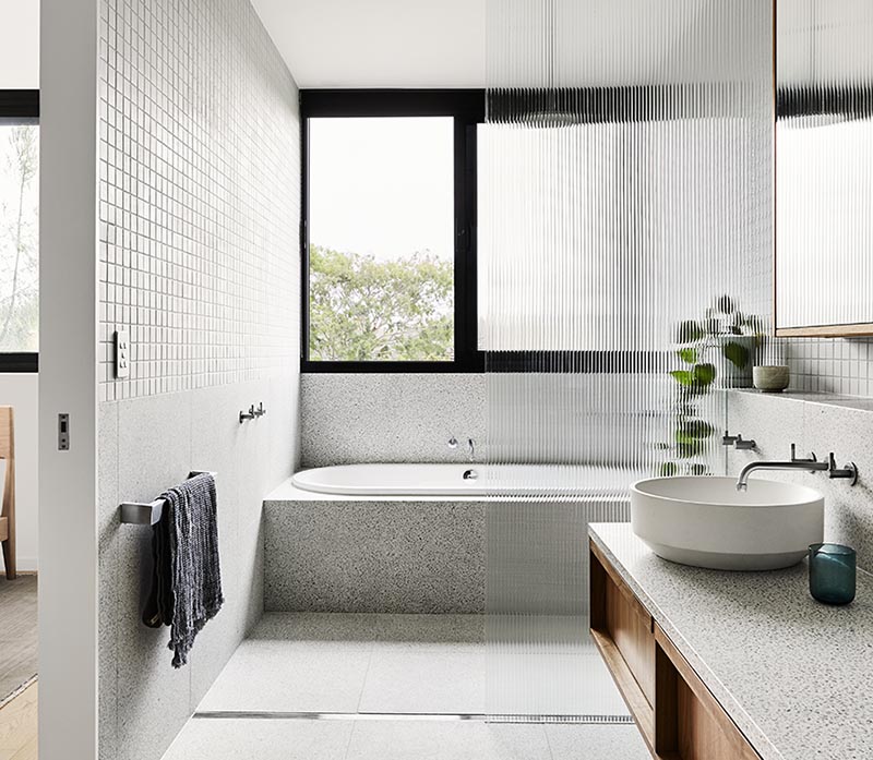 The color palette of this modern bathroom draws inspiration from the surrounding natural landscape found outside the home. The floating wood dual-sink vanity is made from spotted gum timber topped with a terrazzo style textured stone surface. Above the vanity is a tri panel mirror that reflects the light from the window in the bedroom. #ModernBathroom #WoodVanity #FloatingVanity #GreyTiles #Terrazzo #BuiltInBathtub