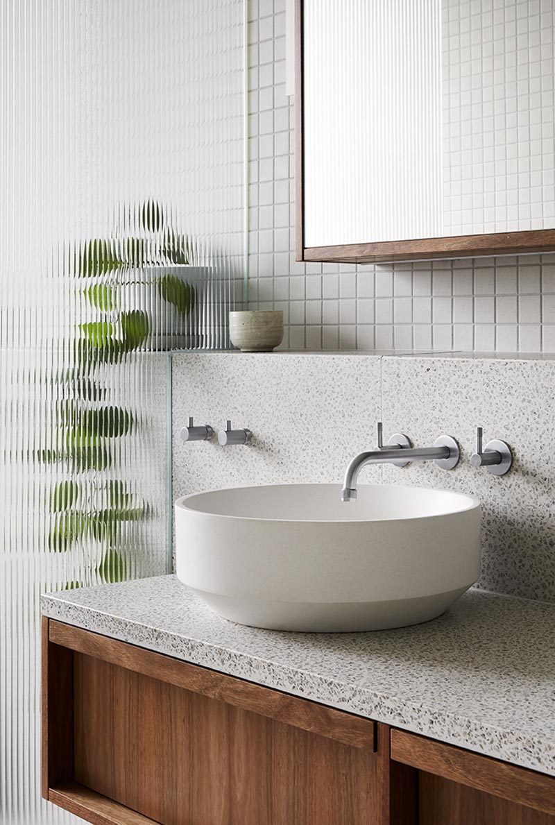 This modern grey bathroom features a glass shower screen that provides a textural element, and above the vanity is small shelf that travels through to the show and creates a place for displaying plants and decorative objects. #ModernBathroom #TexturedShowerScreen #BathroomVanity #Terrazzo #GreyTiles #WoodVanity