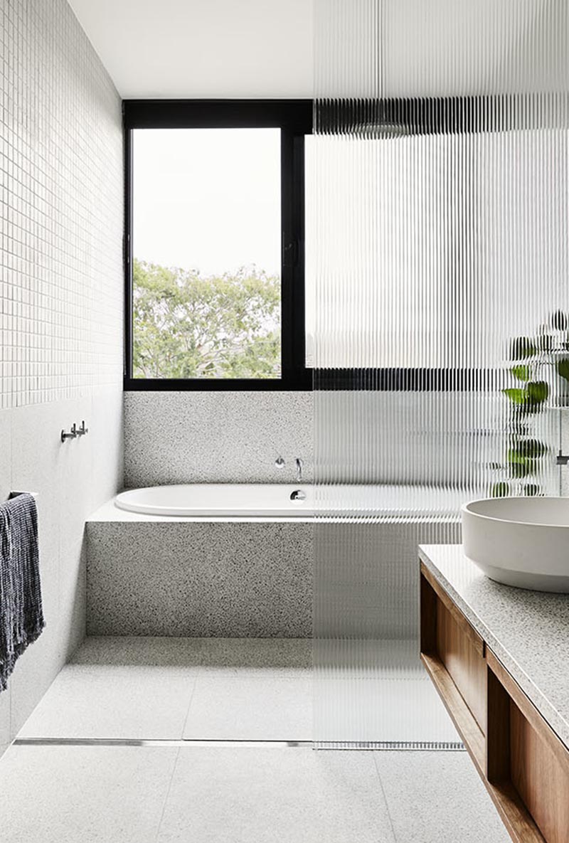 Small grey square tiles cover the upper half of the bathroom walls, while large format terrazzo-style tiles are showcased on the lower portion of the walls and floor, where a linear shower drain can be found. #ModernBathroom #BathroomDesign #BuiltInBathtub #TexturedShowerScreen #GreyBathroom
