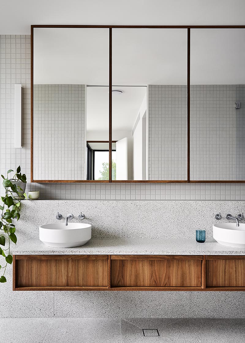 The color palette of this modern bathroom draws inspiration from the surrounding natural landscape found outside the home. The floating wood dual-sink vanity is made from spotted gum timber topped with a terrazzo style textured stone surface. Above the vanity is a tri panel mirror that reflects the light from the window in the bedroom. #ModernBathroom #WoodVanity #FloatingVanity #GreyTiles #Terrazzo