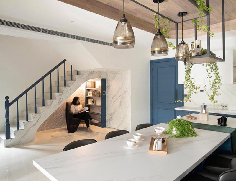 This modern house has a small reading nook under the stairs. It features a grey tile accent wall, a custom blue bookshelf, a comfortable armchair, a sconce, and a pair of outlets. #ReadingNook #SeatingNook #UnderStairNook #HomeLibrary