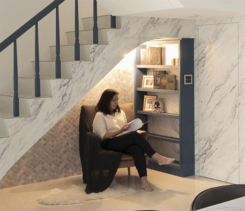 This modern house has a small reading nook under the stairs. It features a grey tile accent wall, a custom blue bookshelf, a comfortable armchair, a sconce, and a pair of outlets. #ReadingNook #SeatingNook #UnderStairNook #HomeLibrary