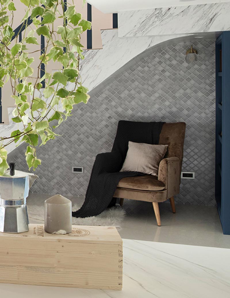 This modern house has a small reading nook under the stairs. It features a grey tile accent wall, a custom blue bookshelf, a comfortable armchair, a sconce, and a pair of outlets. #ReadingNook #SeatingNook #UnderStairNook #HomeLibrary