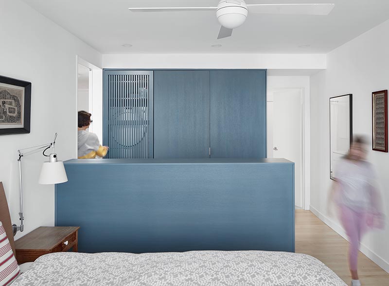 In this modern bedroom, a wall of matte-blue-grey stained closets has one with a wood slat door, which when opened tucks away to the side and reveals a laundry area with a washer / dryer and shelving above. #SmallLaundry #SmallSpaceIdeas #LaundryIdeas #ClosetIdeas