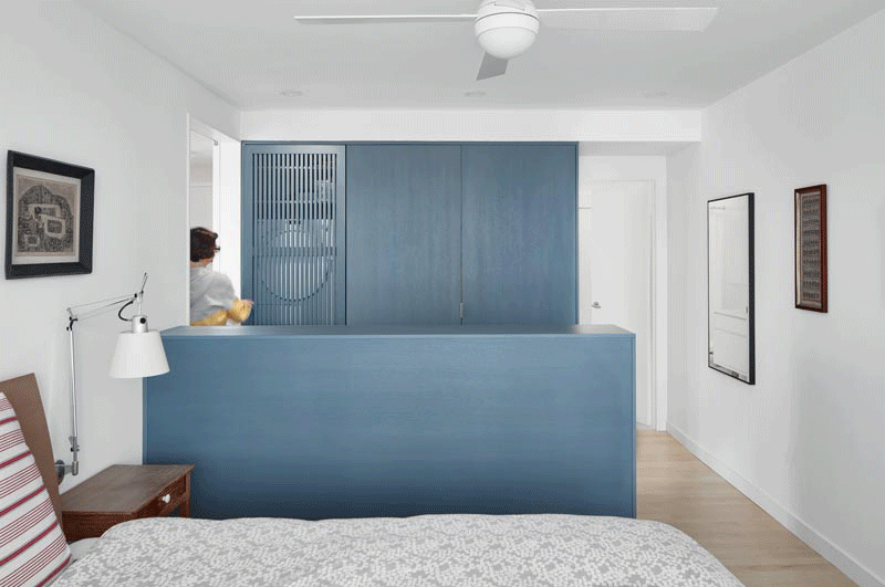 In this modern bedroom, a wall of matte-blue-grey stained closets has one with a wood slat door, which when opened tucks away to the side and reveals a laundry area with a washer / dryer and shelving above. #SmallLaundry #SmallSpaceIdeas #LaundryIdeas #ClosetIdeas