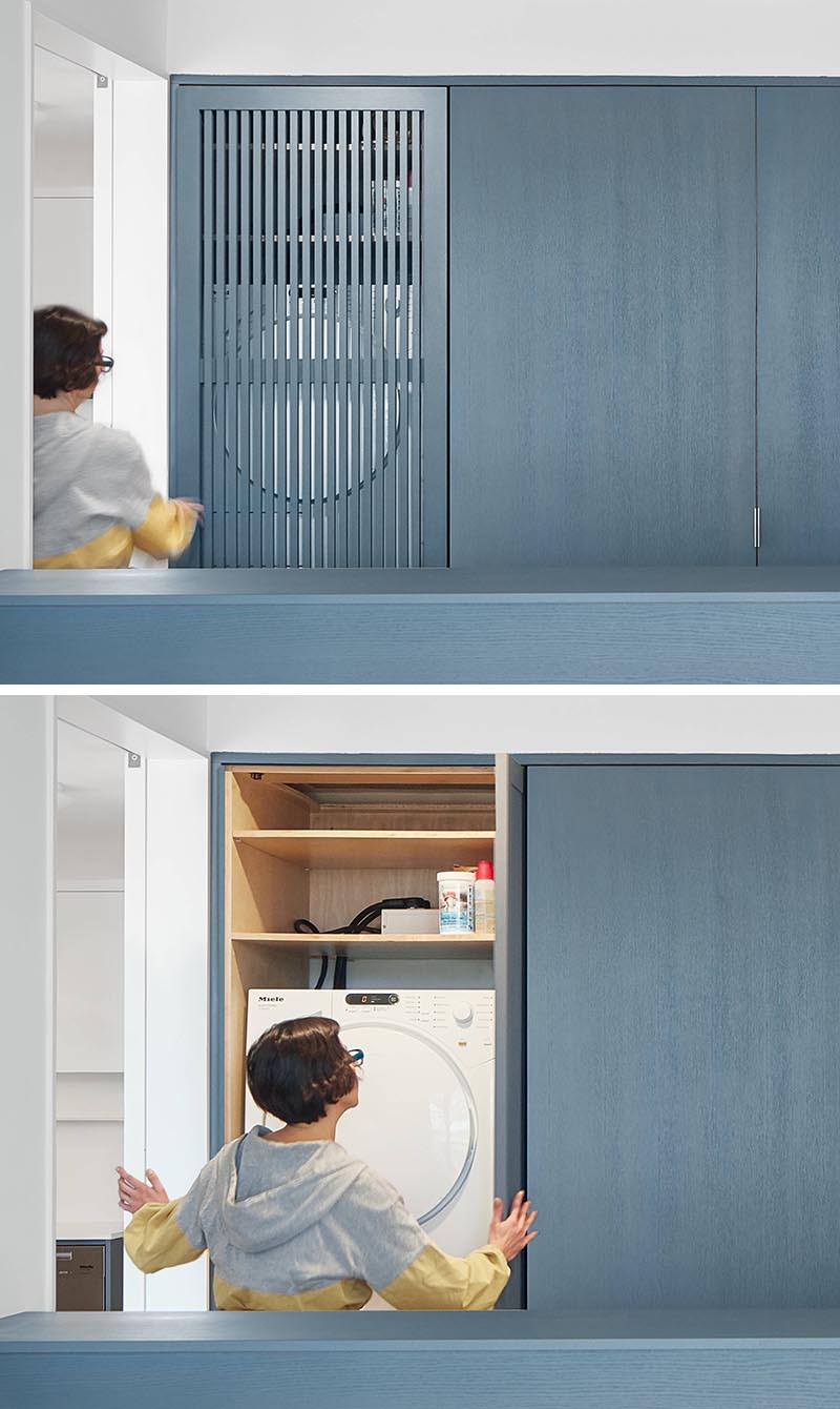 In this modern bedroom, a wall of matte-blue-grey stained closets has one with a wood slat door, which when opened tucks away to the side and reveals a laundry area with a washer / dryer and shelving above. #SmallLaundry #SmallSpaceIdeas #LaundryIdeas #ClosetIdeas