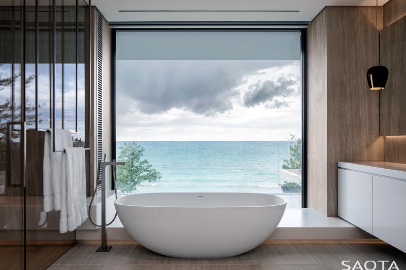 In this modern master bathroom, a freestanding bathtub is located directly in front of a large picture window that provides an uninterrupted view of the lake, and looks like a moving painting. #ModernMasterBathroom #BathroomDesign #FreestandingBathtub #PictureWindow